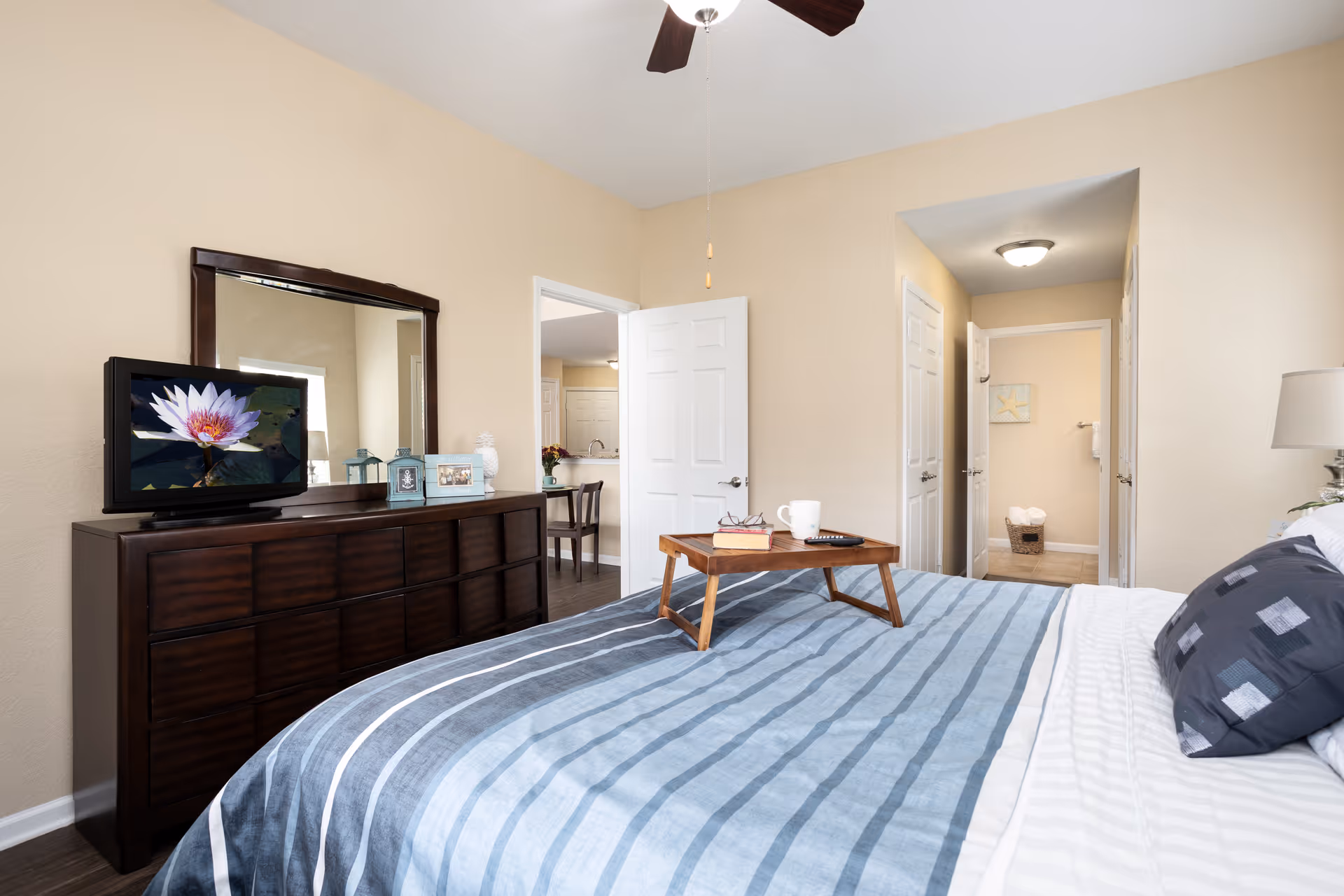 A bright bedroom with a neatly made bed featuring a blue and white striped comforter and a dark pillow. A wooden tray with a book, glasses, a coffee mug, and a remote control rests on the bed. Against the wall is a dark wooden dresser with a large mirror above it and a small TV displaying a flower. The room has beige walls and an open door leading to a hallway and another room.