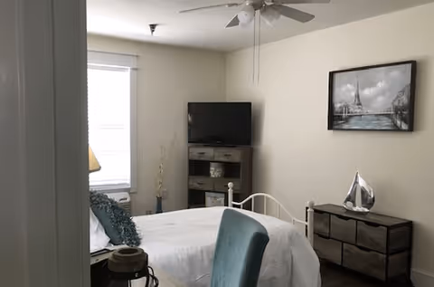 A bedroom with a white bed, teal chair, and a dresser with a decorative silver sailboat on top. A flat-screen TV is on a stand in the corner, and a framed picture of the Eiffel Tower hangs on the wall. The room has a ceiling fan and a window with blinds partially open.