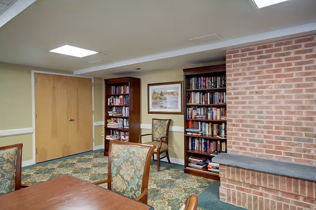 Interior room with a wooden table and upholstered chairs with floral patterns, two wooden bookshelves filled with books, a framed painting on the wall, a double wooden door, and a brick fireplace on the right side.