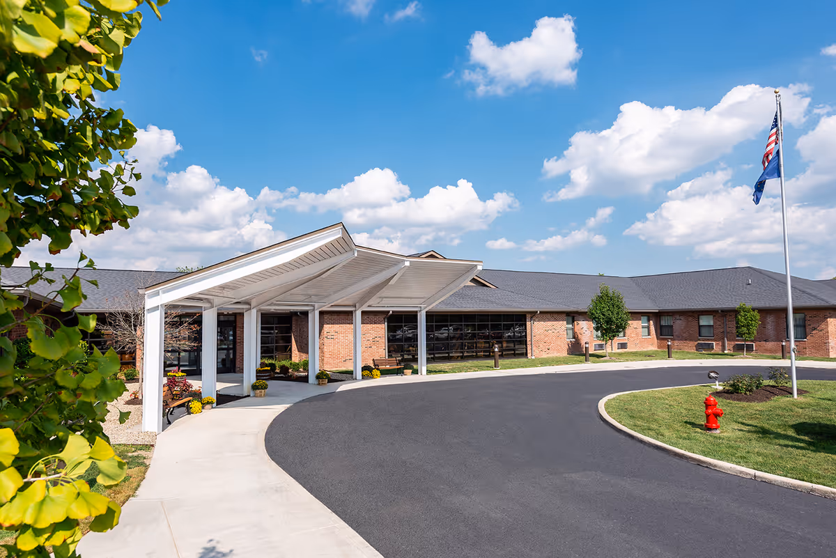 Exterior view of University Heights Health & Living Community building with a covered entrance, a curved driveway, a fire hydrant, an American flag on a flagpole, and a clear blue sky with scattered clouds.