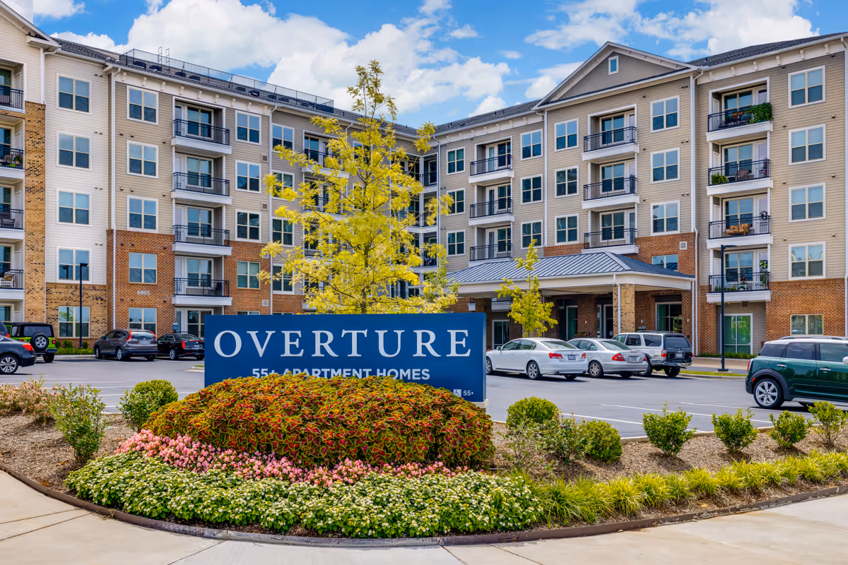 Exterior view of a multi-story residential building with balconies and numerous windows. In front of the building is a landscaped area with colorful flowers and shrubs, and a large blue sign that reads 'OVERTURE 55+ APARTMENT HOMES'. Several cars are parked in the parking lot near the entrance. The sky is partly cloudy with blue patches.
