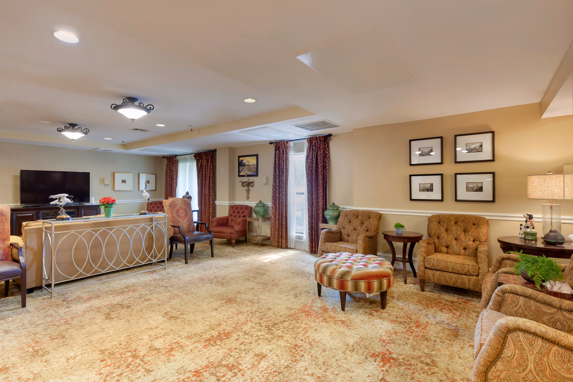 Spacious assisted-living lounge with multiple upholstered chairs and sofas, a patterned ottoman, TV, side tables, and curtains.