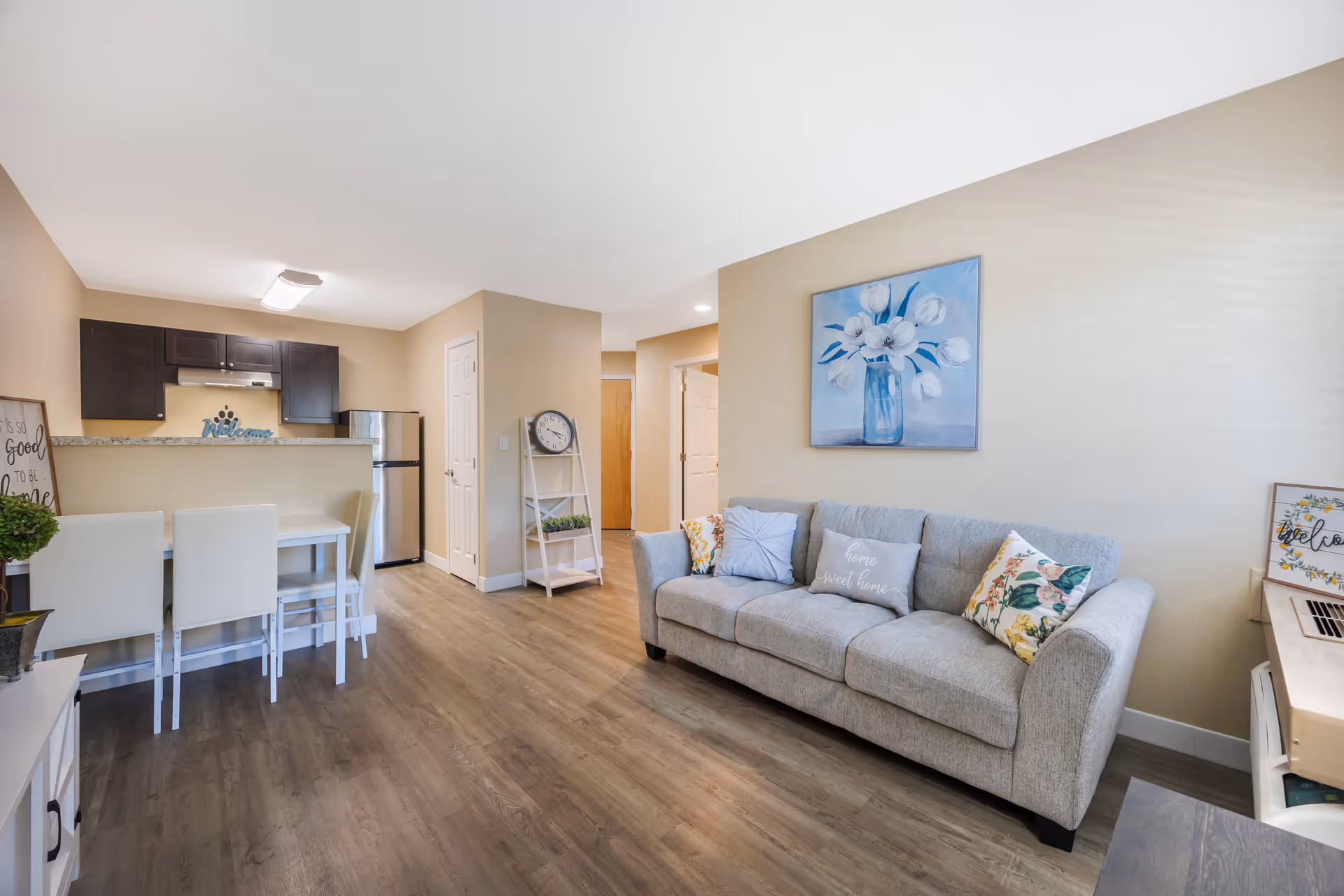 Bright open-plan living area with a gray sofa, floral wall art, and a small kitchen with a breakfast bar and stools.