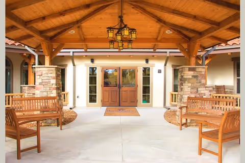 Covered entrance area of a building with wooden benches on either side, stone pillars supporting a wooden roof structure, and double wooden doors with glass panels in the center. A chandelier hangs from the ceiling, and a welcome mat is placed in front of the doors.