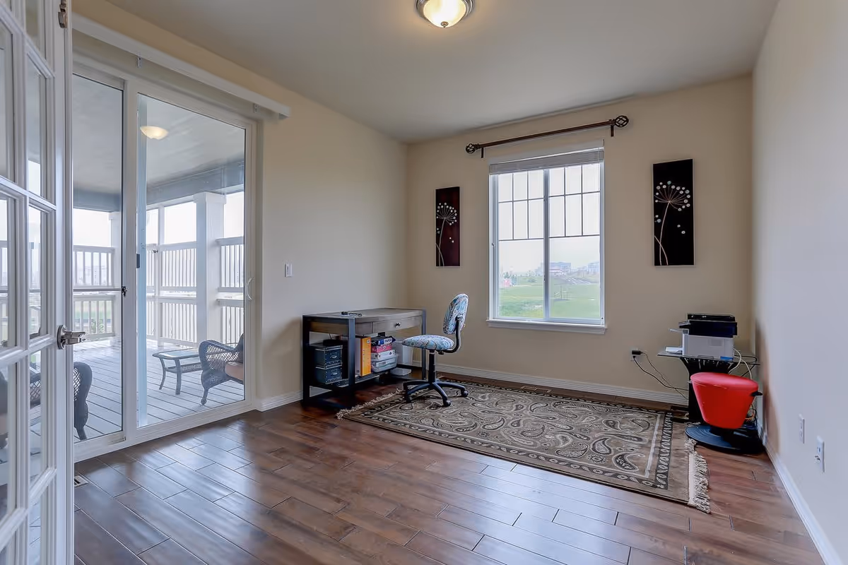 Small bright office with hardwood floors, a desk and rolling chair, patterned rug, and sliding glass doors leading to a covered balcony.