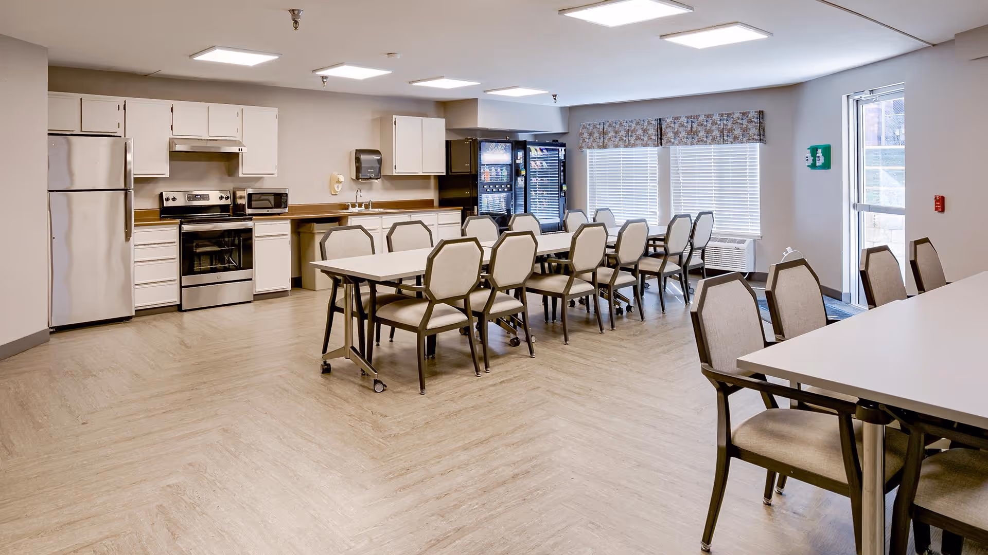 A spacious dining area with multiple tables and chairs arranged neatly. The room features a kitchen area with stainless steel appliances including a refrigerator, oven, and microwave. Large windows with blinds and a patterned valance allow natural light to fill the room. The floor has a light wood finish and the walls are painted in neutral tones.