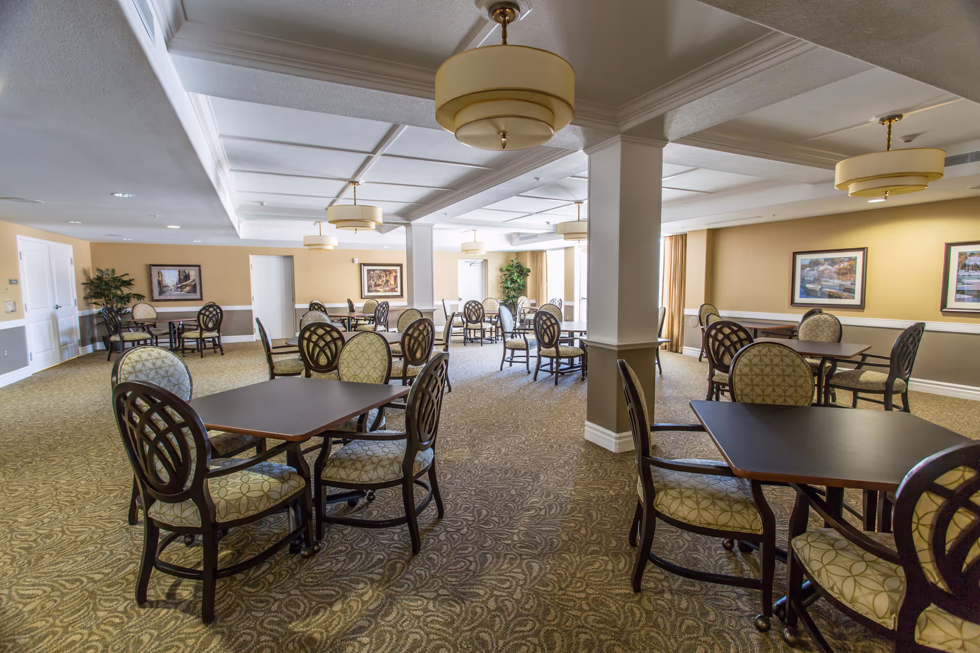 Spacious dining room with multiple square tables and patterned upholstered chairs under pendant lights in a senior living facility.