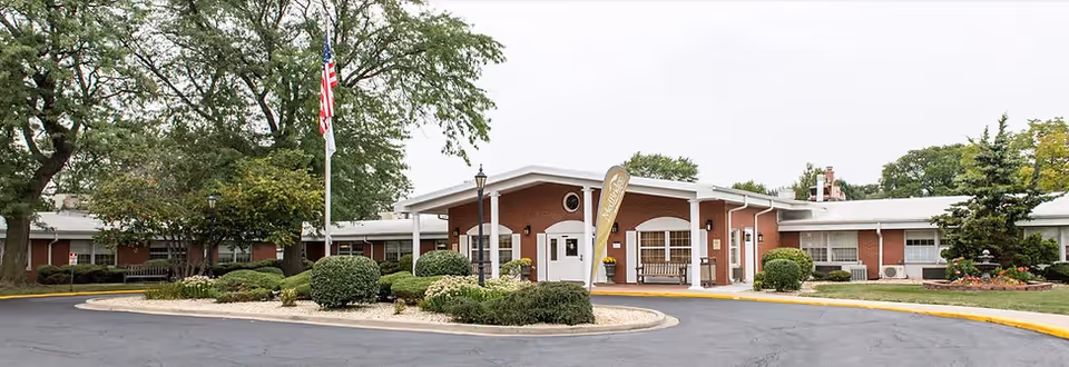 Front exterior view of Manor Care facility showing a single-story brick building with a covered entrance, surrounded by landscaped bushes and trees, an American flag on a flagpole, and a circular driveway.