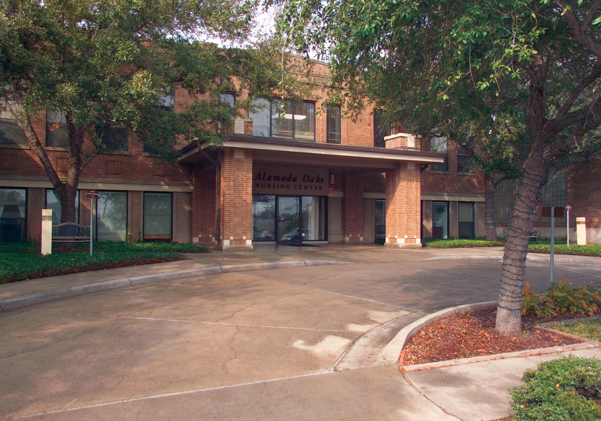 Front exterior view of Alameda Oaks Nursing Center, a two-story brick building with a covered entrance and surrounding trees and landscaping.