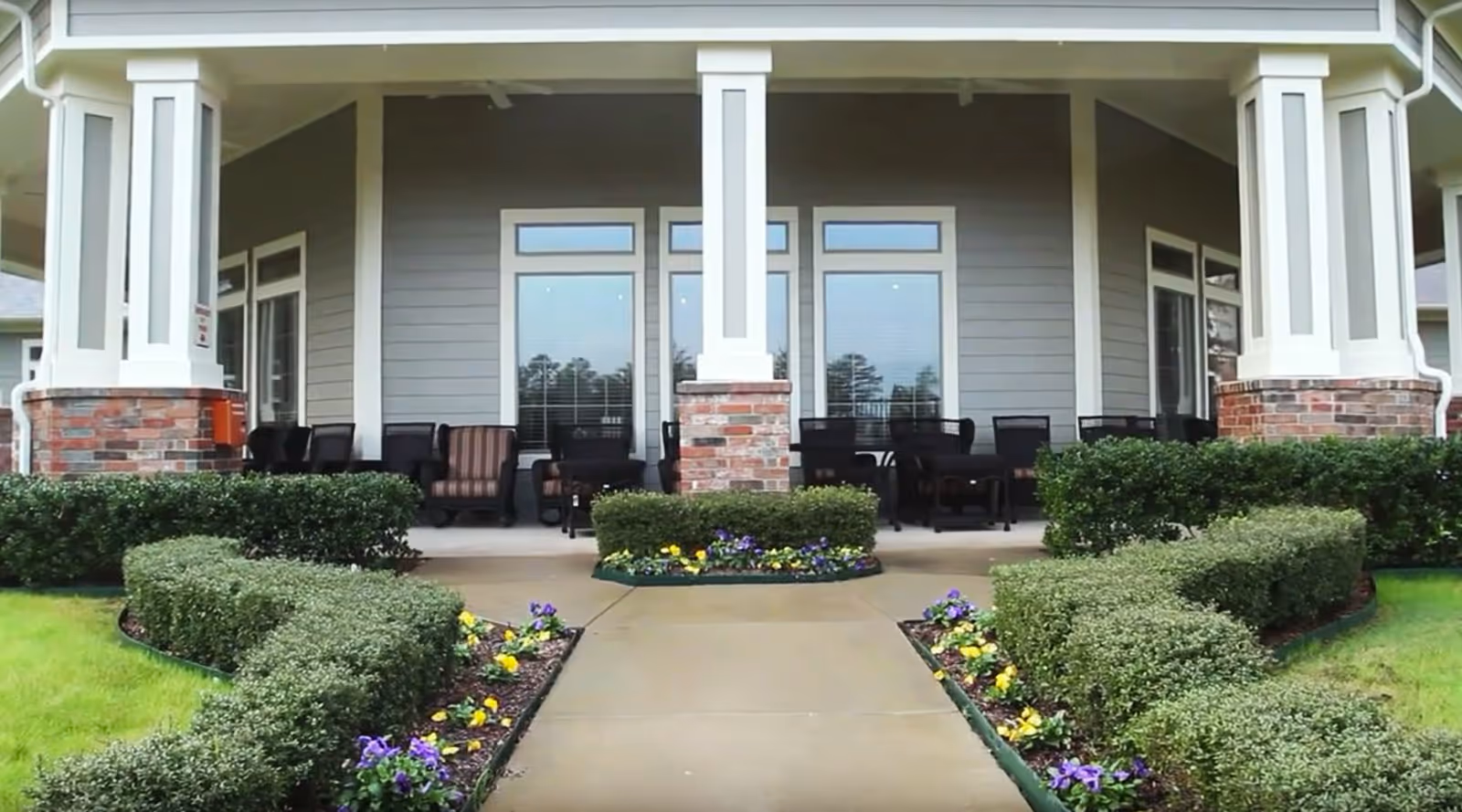 Outdoor covered patio area with multiple black chairs and tables, surrounded by neatly trimmed bushes and flower beds with yellow and purple flowers, in front of a building with gray siding and white trim.