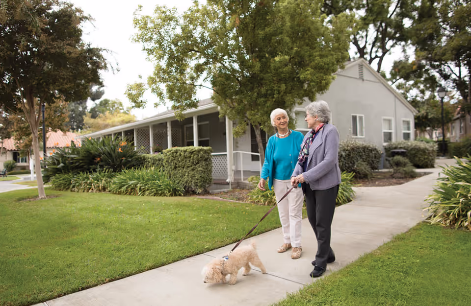 Two elderly women walking a small dog on a leash along a paved pathway in a green, landscaped outdoor area of a retirement community with single-story buildings and trees in the background.