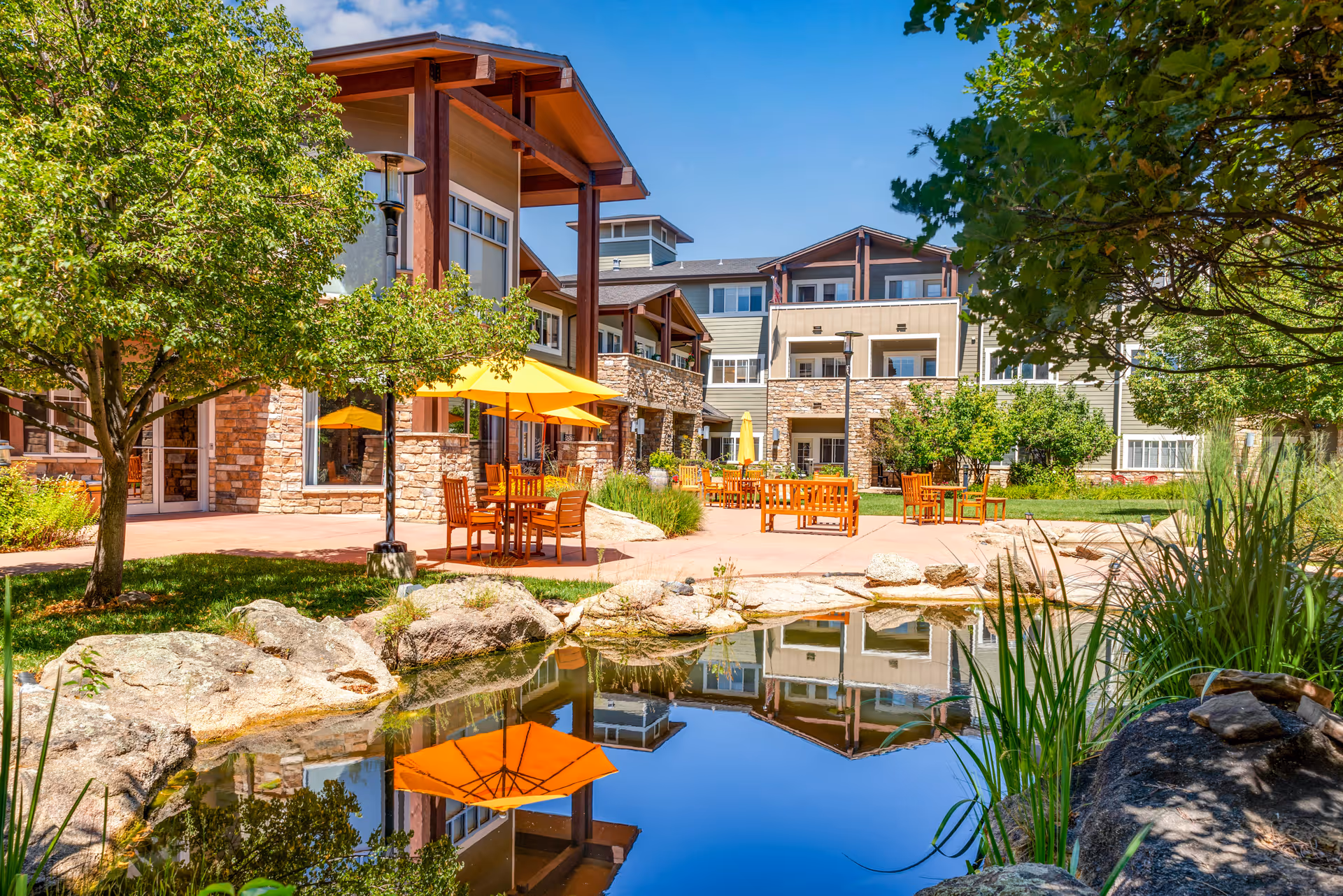Outdoor patio area at Balfour Louisville Campus featuring wooden tables and chairs with yellow umbrellas, surrounded by trees and greenery. A small pond with rocks and plants reflects the building and umbrellas. The multi-story building has stone and wood accents under a clear blue sky.