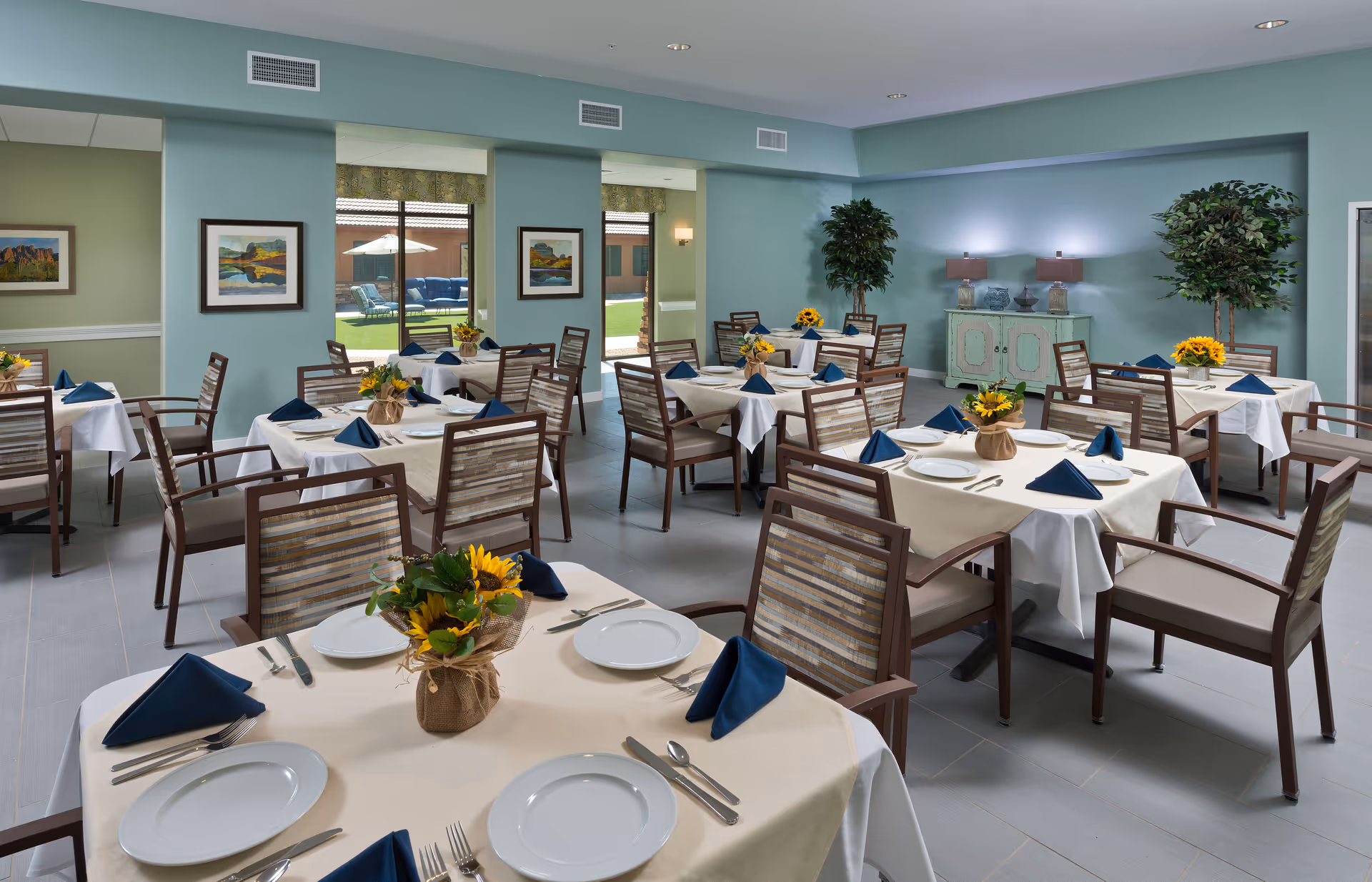 Dining room with multiple tables set with white tablecloths, blue napkins, plates and sunflower centerpieces.