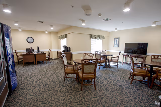 Community dining/activity room with round tables and chairs, a TV, vending machine, coffee station and desks.