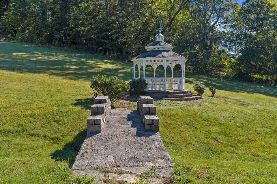 A white wooden gazebo with a shingled roof situated on a grassy hill, surrounded by trees and bushes. A stone pathway with low stone walls on either side leads up to the gazebo.