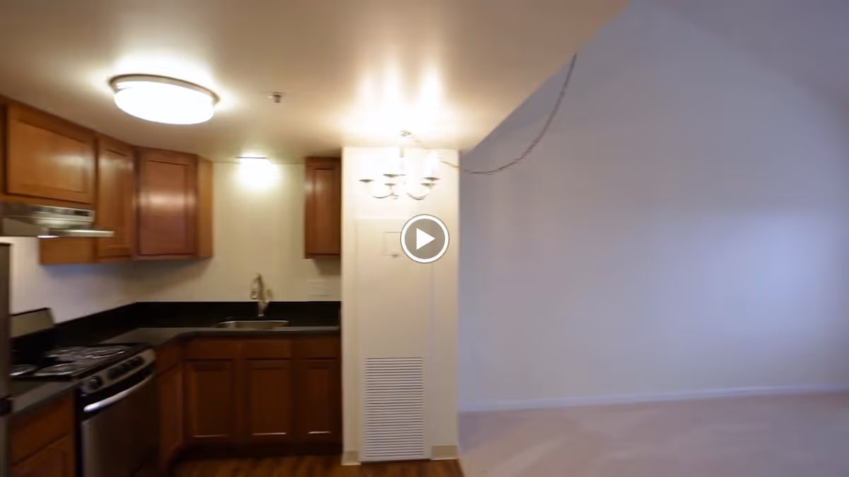 Interior view of a kitchen area with wooden cabinets, a stainless steel stove and oven, a sink, and black countertops. Adjacent to the kitchen is an empty room with beige carpet and white walls, illuminated by a ceiling light fixture.