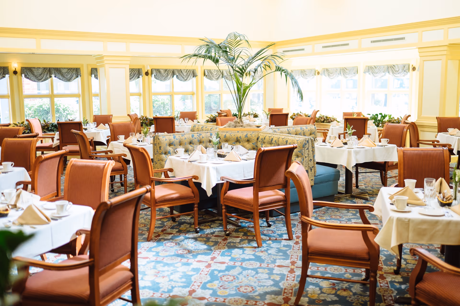 A bright and elegant dining room with multiple tables set with white tablecloths, napkins, cups, and glassware. The room features wooden chairs with orange upholstery, a patterned carpet, large windows with valances, and a central decorative plant arrangement.