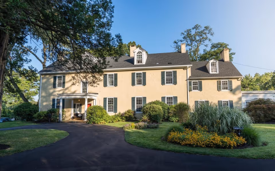 Exterior view of a large, two-story beige building with green shutters and a dark roof, surrounded by well-maintained landscaping including bushes, trees, and flower beds, under a clear blue sky.