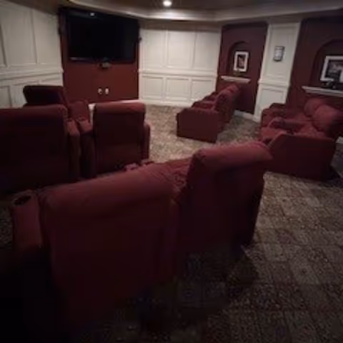 A cozy room with multiple maroon recliner chairs arranged in rows facing a wall-mounted flat screen TV. The walls are decorated with white paneling and maroon paint, and there are framed pictures on the walls. The floor is carpeted with a patterned design.