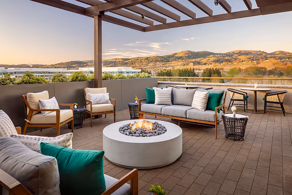 Outdoor patio area with comfortable seating including cushioned chairs and a sofa around a circular fire pit, small tables with flowers, and a pergola overhead. The background shows a scenic view of hills and trees under a clear sky during sunset.