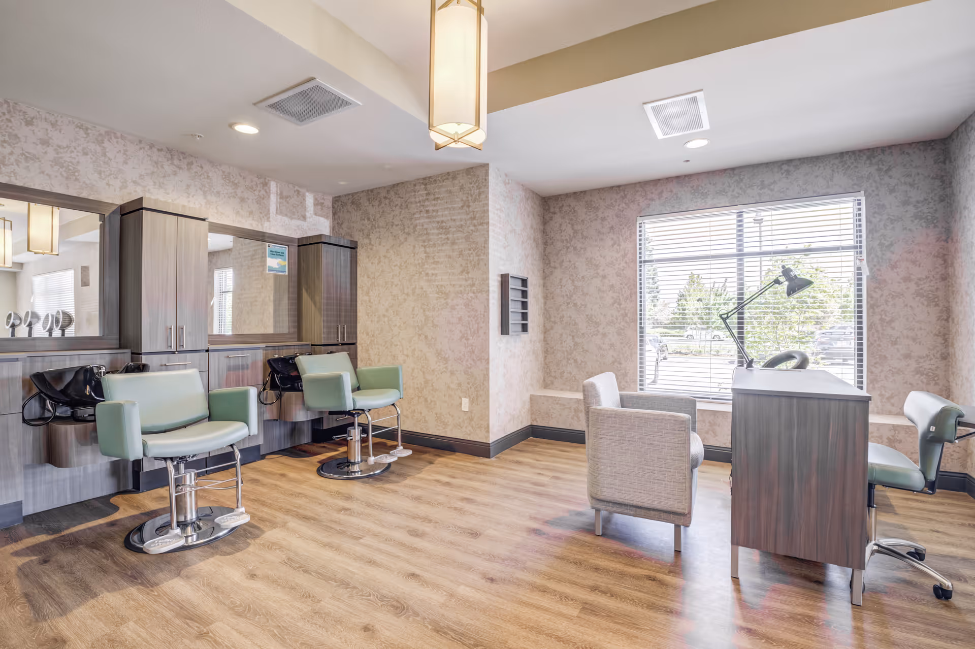 A bright and clean salon area in a senior living facility with two green salon chairs in front of mirrors and cabinetry on the left side, and a small desk with two chairs near a large window with blinds on the right side. The room has wood flooring and soft beige patterned walls.