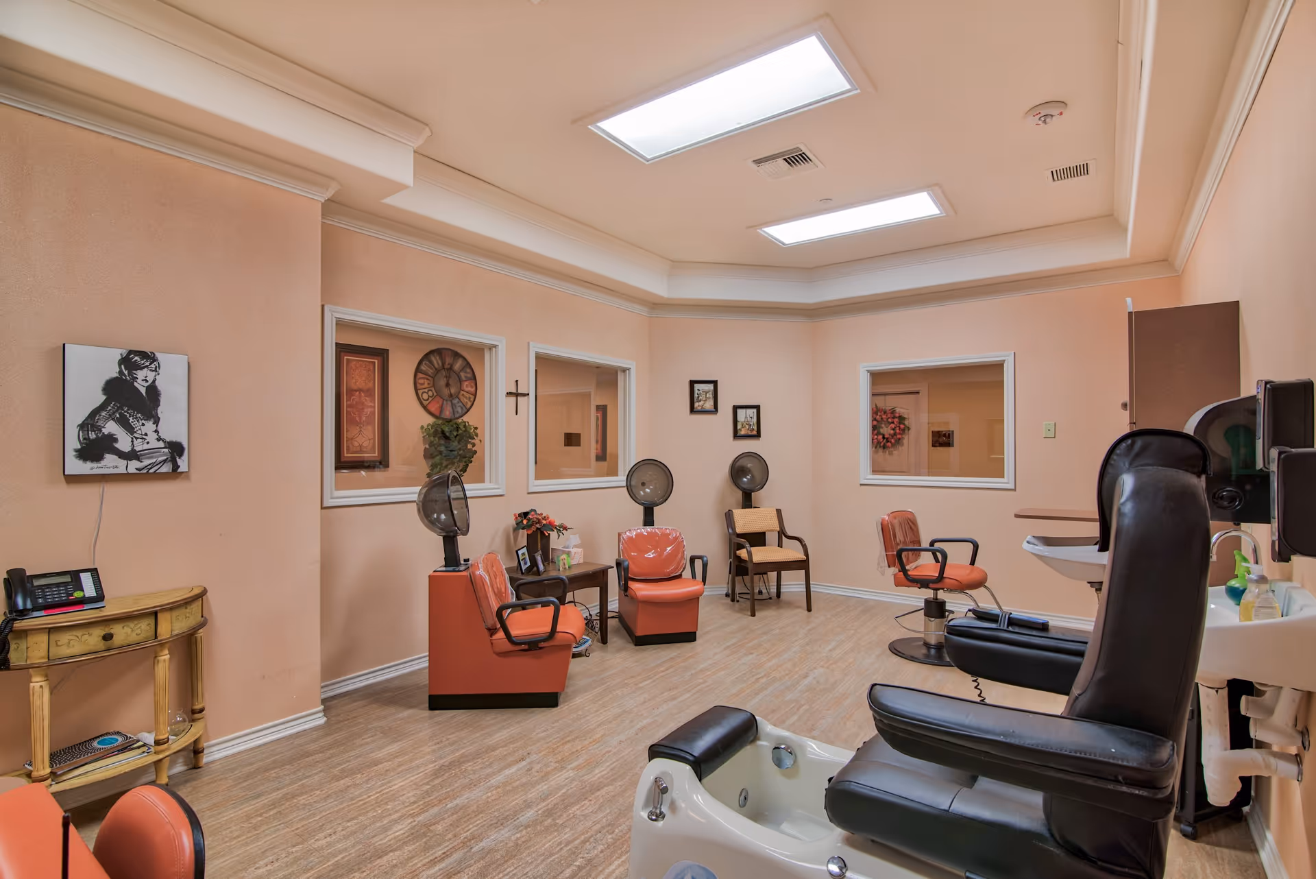 A bright salon-style room in an assisted living facility with pedicure stations, salon chairs and hooded hair dryers against peach-colored walls.