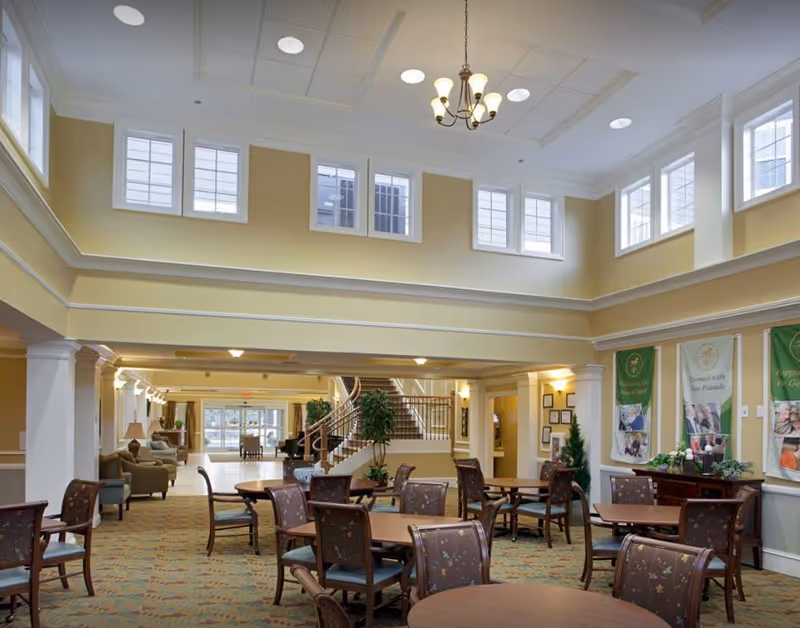 Spacious communal dining and seating area with multiple tables and chairs, high ceilings, many windows, and a central staircase in the background.