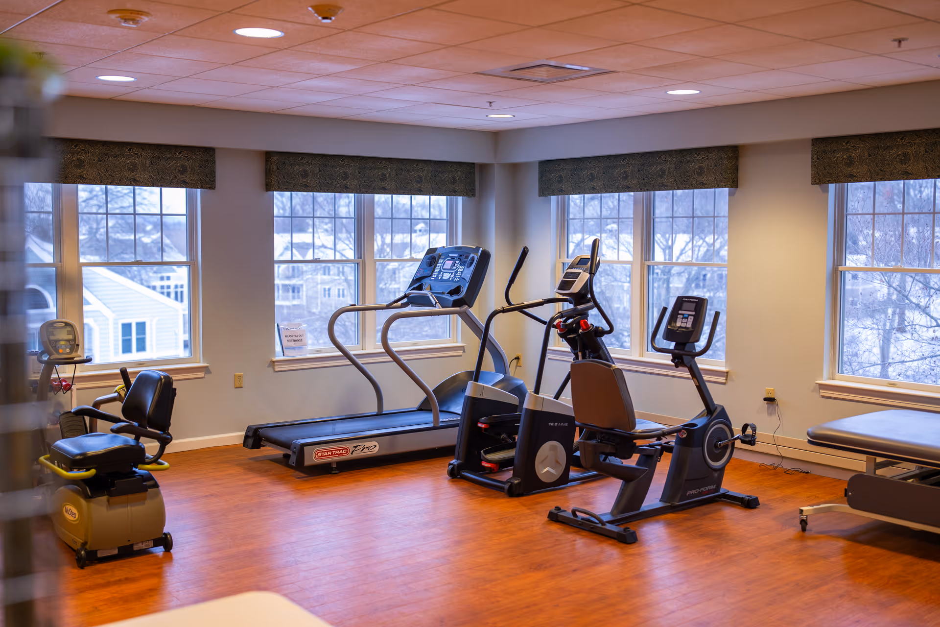 Bright fitness room with treadmills, stationary bikes and exercise equipment arranged on a wood floor in front of large windows.