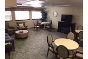 A common area in a senior living facility with multiple round tables and chairs, a flat-screen TV on a stand, a clock on the wall, a large window with blinds, and a green carpeted floor. There is also a green armchair and a round ottoman near the window.