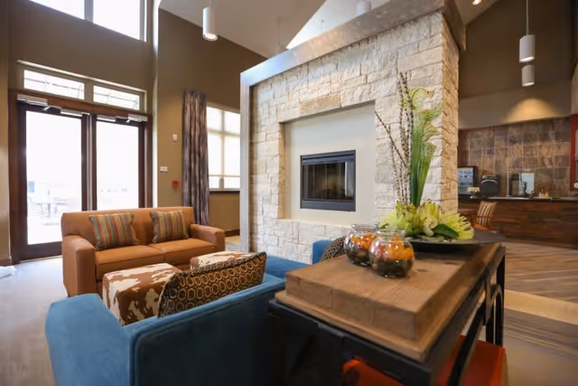 A cozy living room area featuring a stone fireplace in the center, surrounded by a blue sofa and a brown sofa with patterned cushions. Large windows and glass doors allow natural light to fill the space. A wooden table with decorative plants and glass bowls is in the foreground, and a kitchenette area is visible in the background.