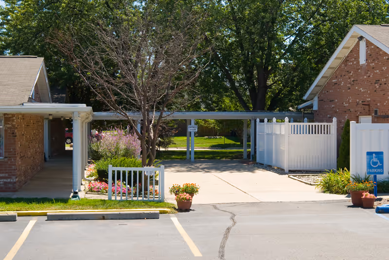 Exterior view of a senior living facility with brick buildings, a covered walkway, a small garden with flowers, a tree without leaves, and a parking area including a handicapped parking sign.