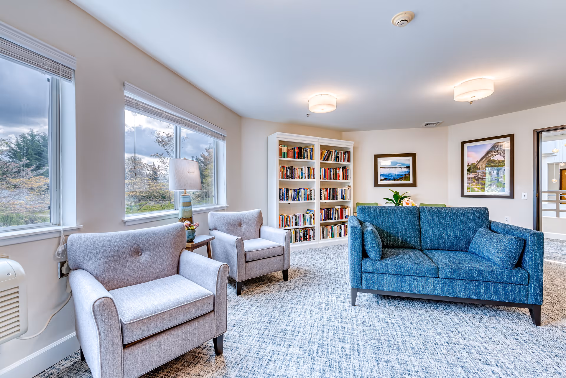 A bright and cozy living room area in a senior living facility featuring two light gray armchairs and a blue sofa arranged around a small wooden side table with a lamp. Behind the seating is a white bookshelf filled with books, and the walls are decorated with framed landscape photographs. Large windows let in natural light and show trees outside.