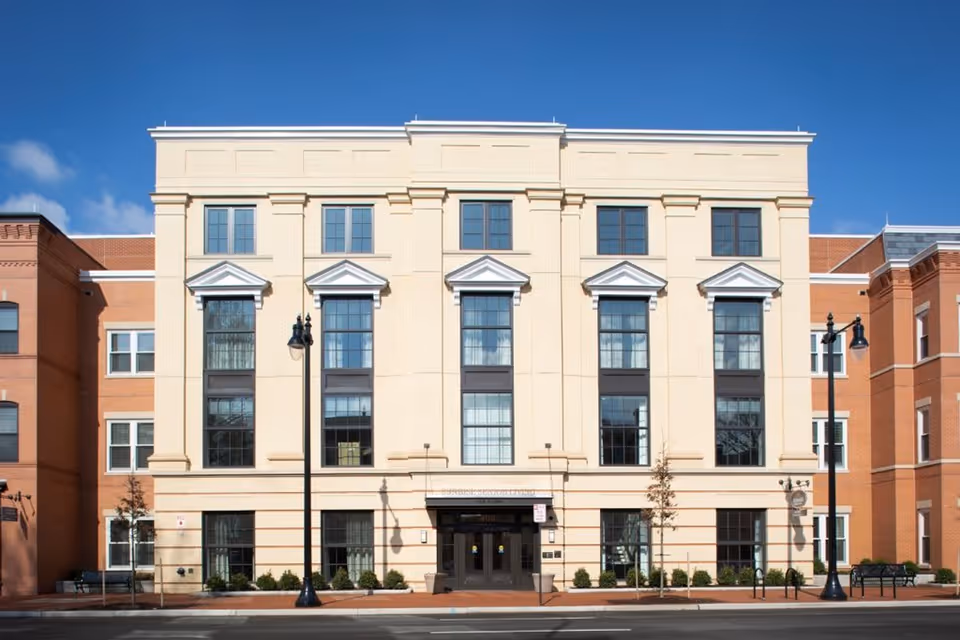 Front exterior view of a multi-story senior living facility building with large windows, beige and brick facade, street lamps, small trees, and benches along the sidewalk under a clear blue sky.
