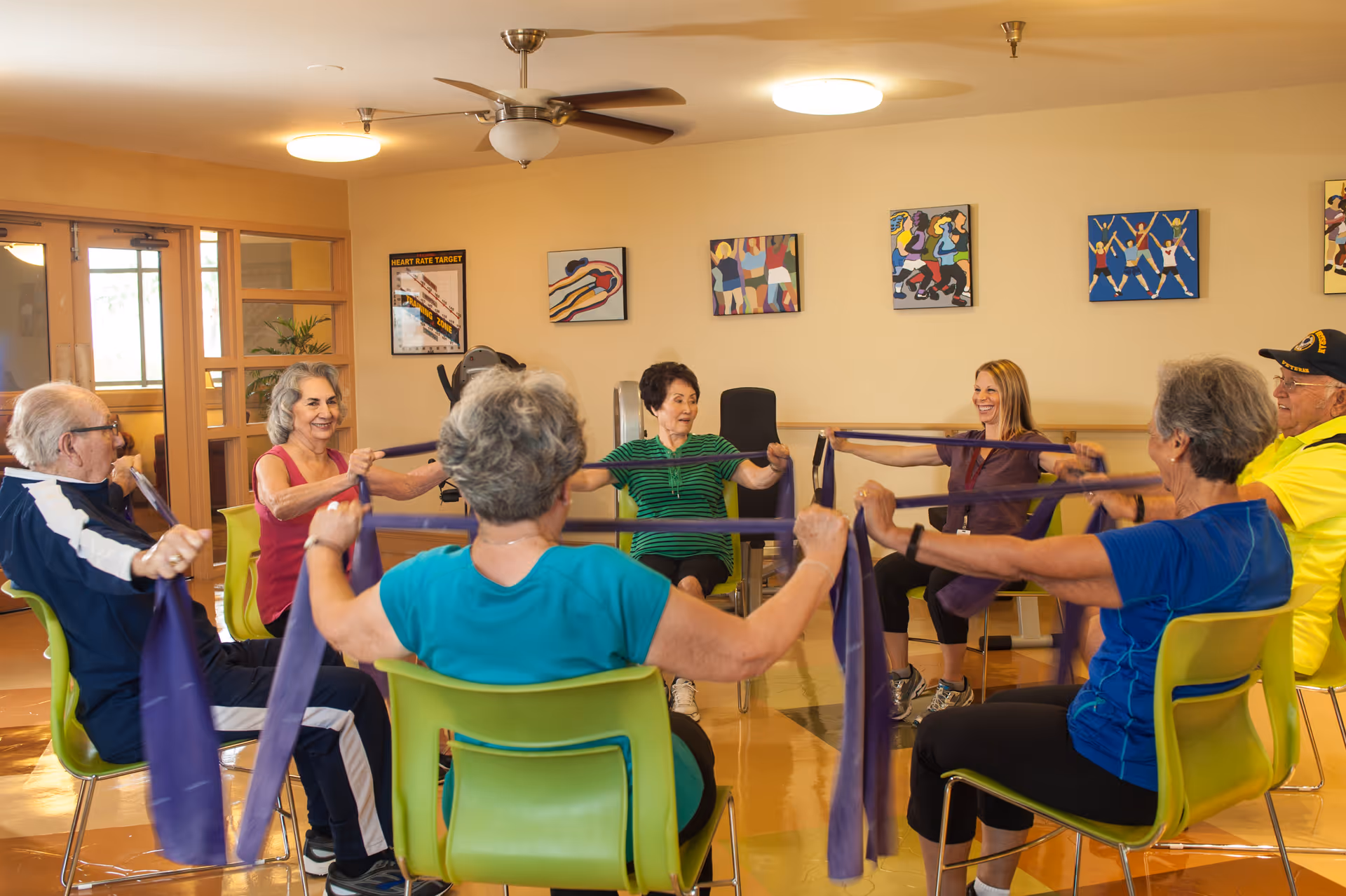 A group of elderly people sitting in a circle on green chairs in a well-lit room, participating in a seated exercise class using purple resistance bands. The room has light yellow walls decorated with colorful artwork and a ceiling fan.
