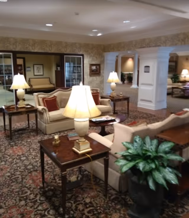 A cozy and elegant living room area in a senior living facility with beige sofas, wooden side tables with lamps, a patterned carpet, and a large potted plant. The room features warm lighting and decorative wallpaper, with an open view into a hallway and additional seating areas.