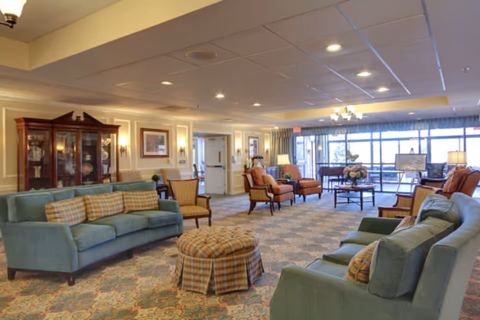 A spacious and well-lit senior living facility common area with comfortable seating including teal sofas, plaid cushions, armchairs, and a round ottoman. The room features a patterned carpet, a wooden display cabinet with glass doors, framed artwork on the walls, and large windows letting in natural light. There are lamps and a chandelier providing additional lighting.