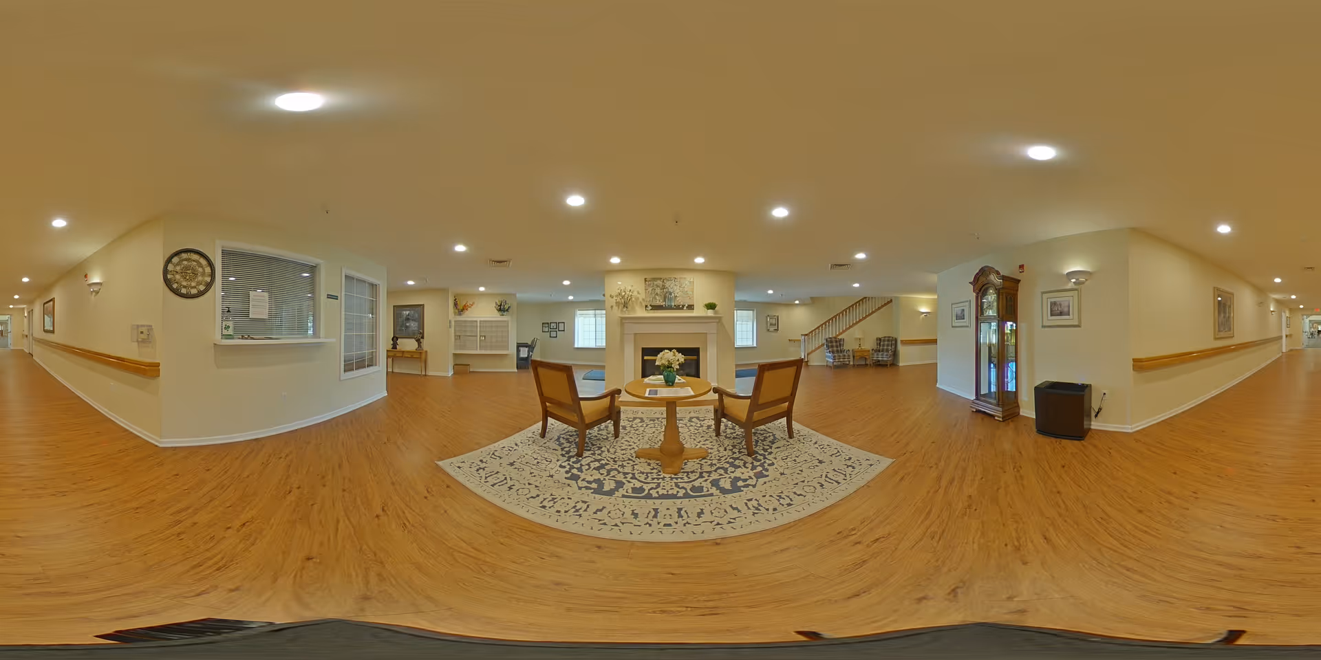A spacious, well-lit common area in an assisted living facility with wooden flooring, a round table with two chairs on a patterned rug, a fireplace, a grandfather clock, and hallways extending on both sides. The walls are painted light beige, and there are framed pictures and wall sconces for lighting.