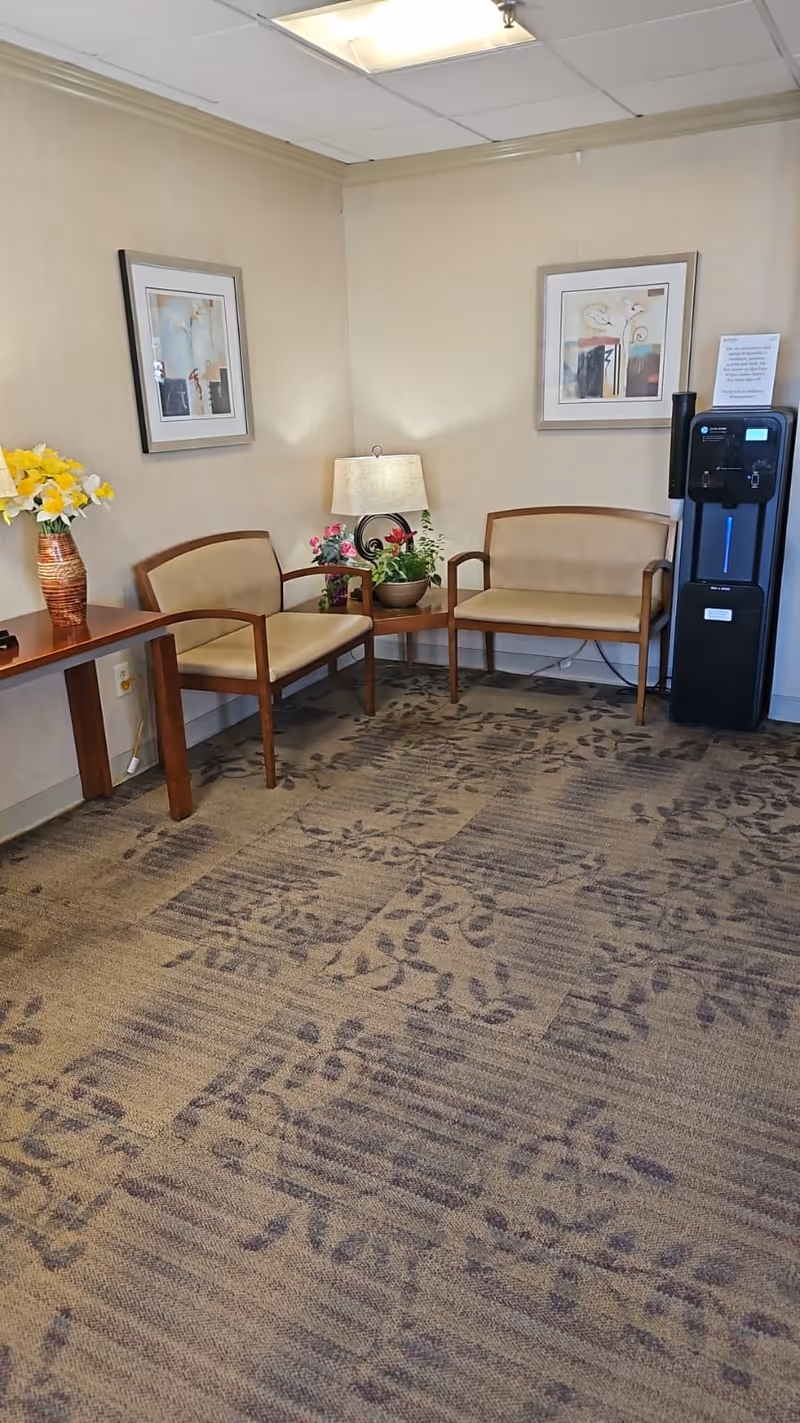A small waiting area with two beige cushioned chairs with wooden frames, a wooden table with a vase of yellow flowers, a small round table with a lamp and a potted plant, two framed abstract paintings on the walls, and a black water cooler in the corner.
