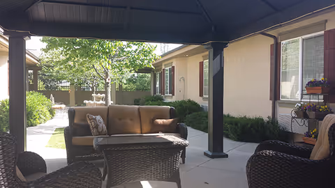 Outdoor covered patio area with wicker furniture including a sofa, chairs, and a table. The patio overlooks a garden with a tree and a decorative water fountain. The building exterior is visible with beige walls and windows with red shutters. Flower pots with colorful flowers are placed on a metal stand near the window.