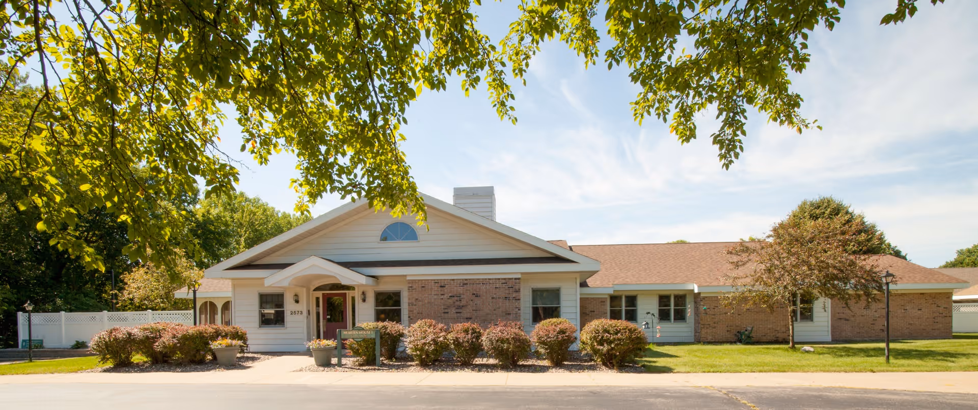 Front exterior view of a single-story senior living facility building with a combination of brick and white siding, a peaked roof, and a covered entrance. The building is surrounded by neatly trimmed bushes, a tree with green leaves in the foreground, and a clear blue sky with some clouds.