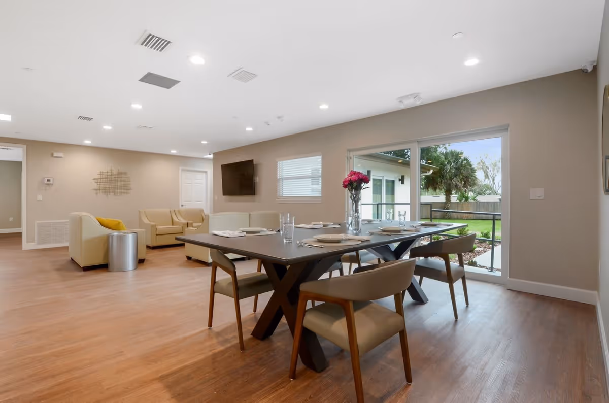 A bright and modern senior living common area featuring a dining table set with plates, glasses, and a vase of pink flowers. The room has beige walls, wood flooring, comfortable beige armchairs and sofas, a wall-mounted TV, and large sliding glass doors that open to a green outdoor space with trees and a fence.
