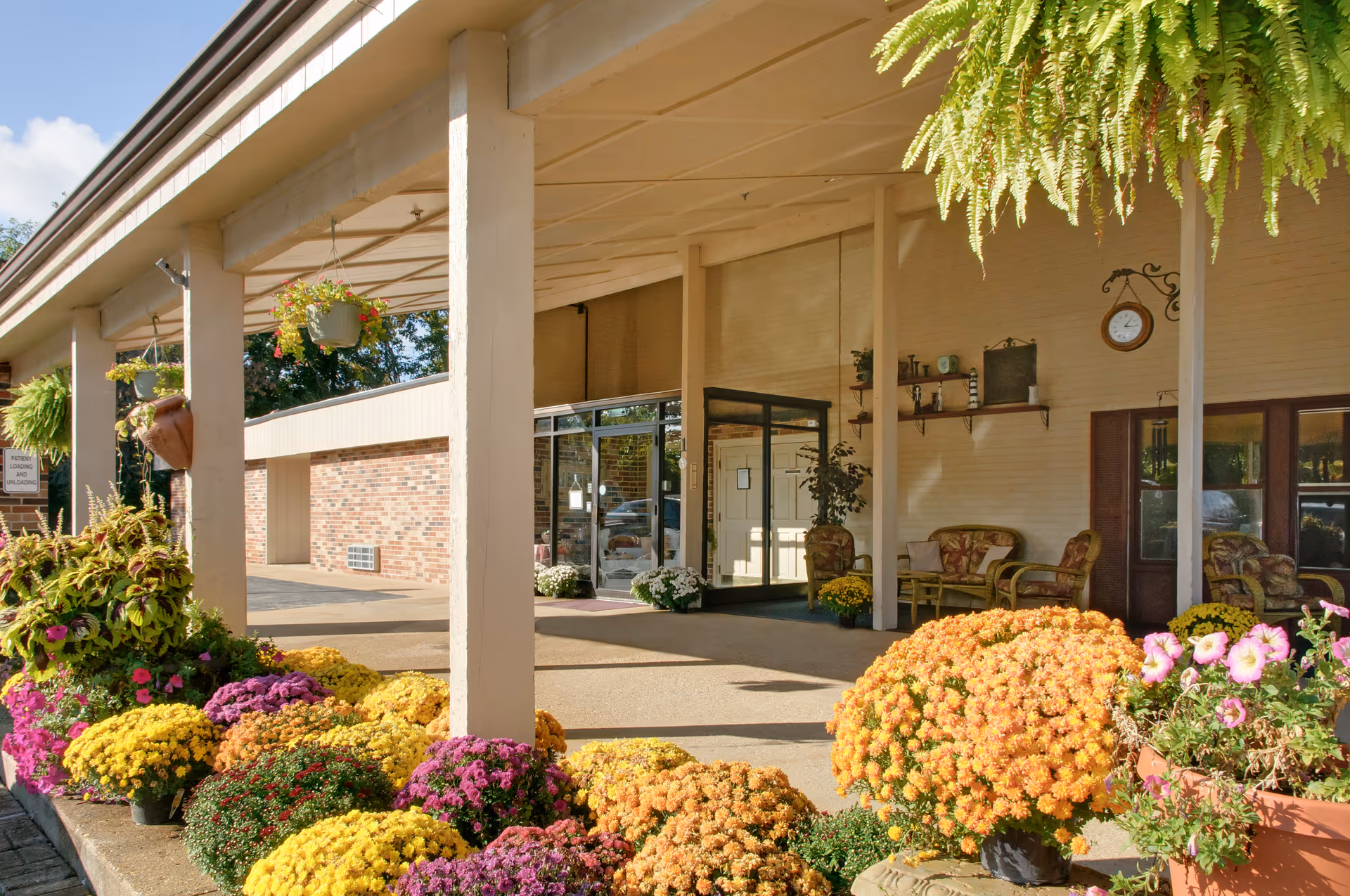 Entrance area of a facility with a covered walkway supported by beige columns. There are numerous colorful potted flowers including yellow, orange, purple, and pink blooms arranged along the walkway. Hanging plants are suspended from the ceiling. The entrance features glass doors and a seating area with cushioned chairs and a small table. A clock and decorative shelves are mounted on the wall near the seating area.
