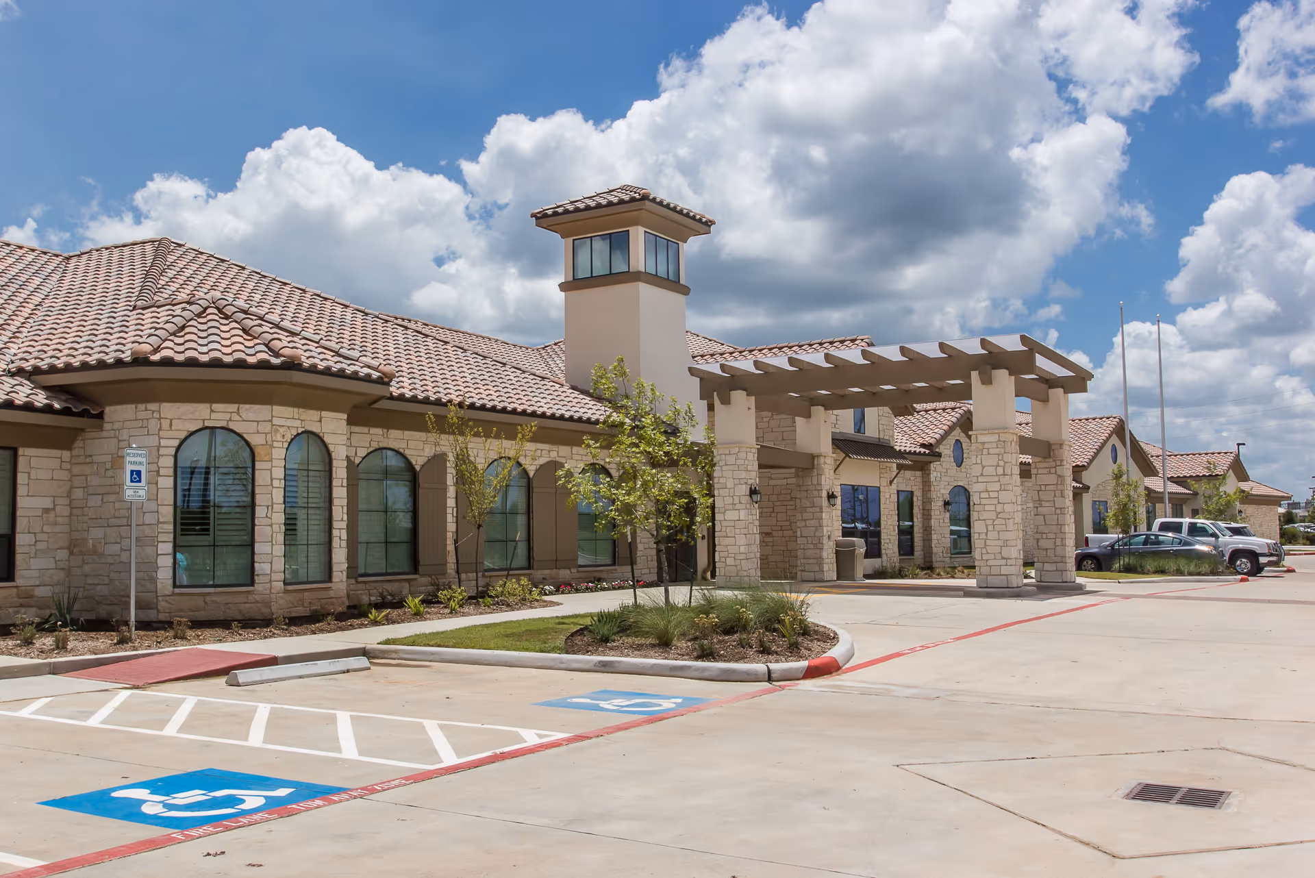 Exterior view of VibraLife El Paso now Ignite Medical Resorts building with stone walls, arched windows, a tiled roof, and a covered entrance. There are handicap parking spaces and a few parked vehicles in the parking lot under a partly cloudy sky.