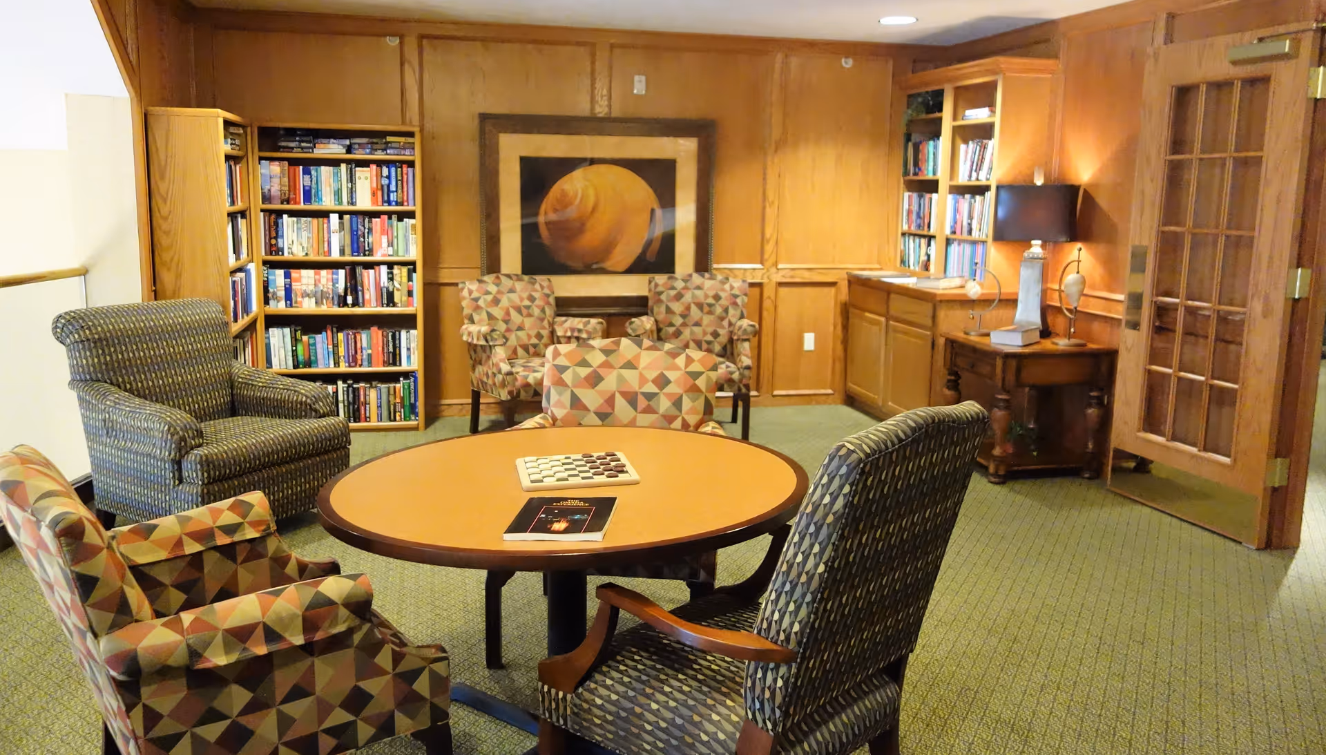 A cozy reading and sitting area in a retirement community featuring a round table with a book and a board game on it, surrounded by four upholstered armchairs with patterned fabric. The room has wood-paneled walls, built-in bookshelves filled with books, a table lamp on a side table, and a framed artwork on the wall. A wooden door with glass panels is visible on the right side.