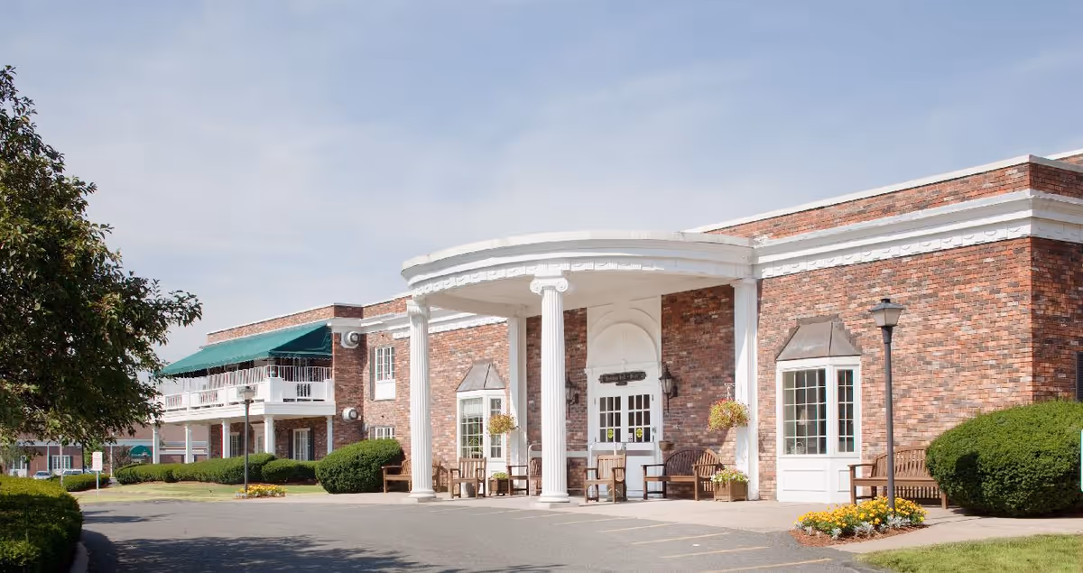 Brick exterior and main entrance of a senior living facility featuring white columns, benches, and landscaped grounds.
