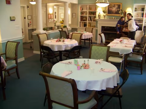 Dining room with round tables draped in white tablecloths, chairs, and a fireplace with bookshelves in the background.