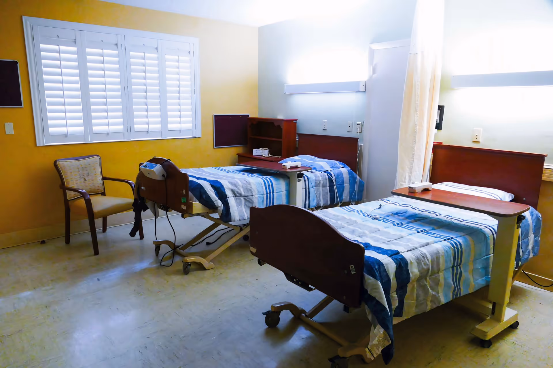 A room with two hospital-style beds with blue and white striped bedding, each equipped with an overbed table. There is a wooden chair near a window with white shutters, and a yellow wall in the background. The room has a clean and simple appearance with medical equipment attached to one of the beds.