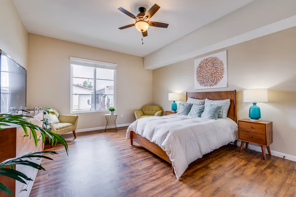 Spacious bedroom with a wooden bed, bedside tables and teal lamps, a window seating area, and hardwood floors.