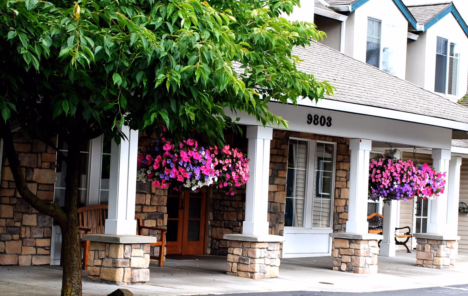 Front exterior view of a senior living facility with stone pillars, white columns, hanging flower baskets with pink, purple, and white flowers, a tree with green leaves, and the address number 9803 displayed above the entrance.
