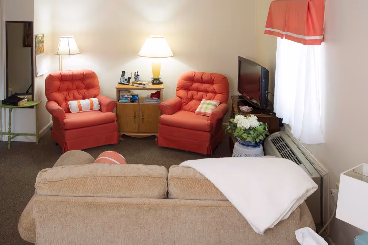 A cozy living room with two coral-colored armchairs, each with a decorative pillow, positioned around a small wooden cabinet with a lamp and some items on top. In front of the chairs is a beige sofa with a white blanket draped over the back. A TV sits on a stand near a window with white curtains and a coral valance. A potted plant is placed next to the TV stand, and a small green side table with a mirror above it is visible on the left side of the room.